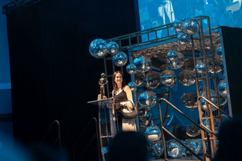 Ania Wysocka at a podium holding an award at an event stage decorated with silver mirror balls