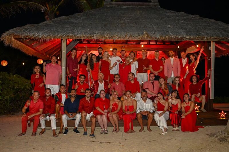 Group of guests in red and white outfits gathered under a thatched beach pavilion at night, holding champagne glasses