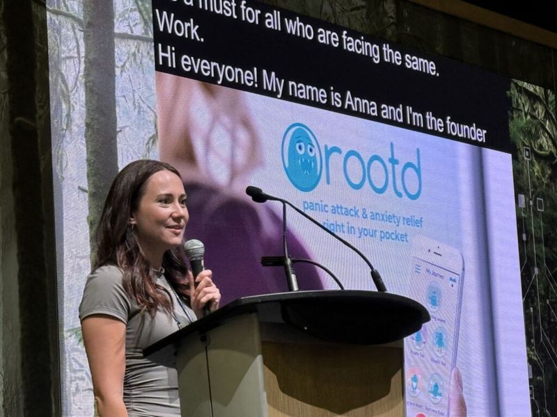 Ania Wysocka speaking at a podium with a microphone, a Rootd app slide projected on screen behind her