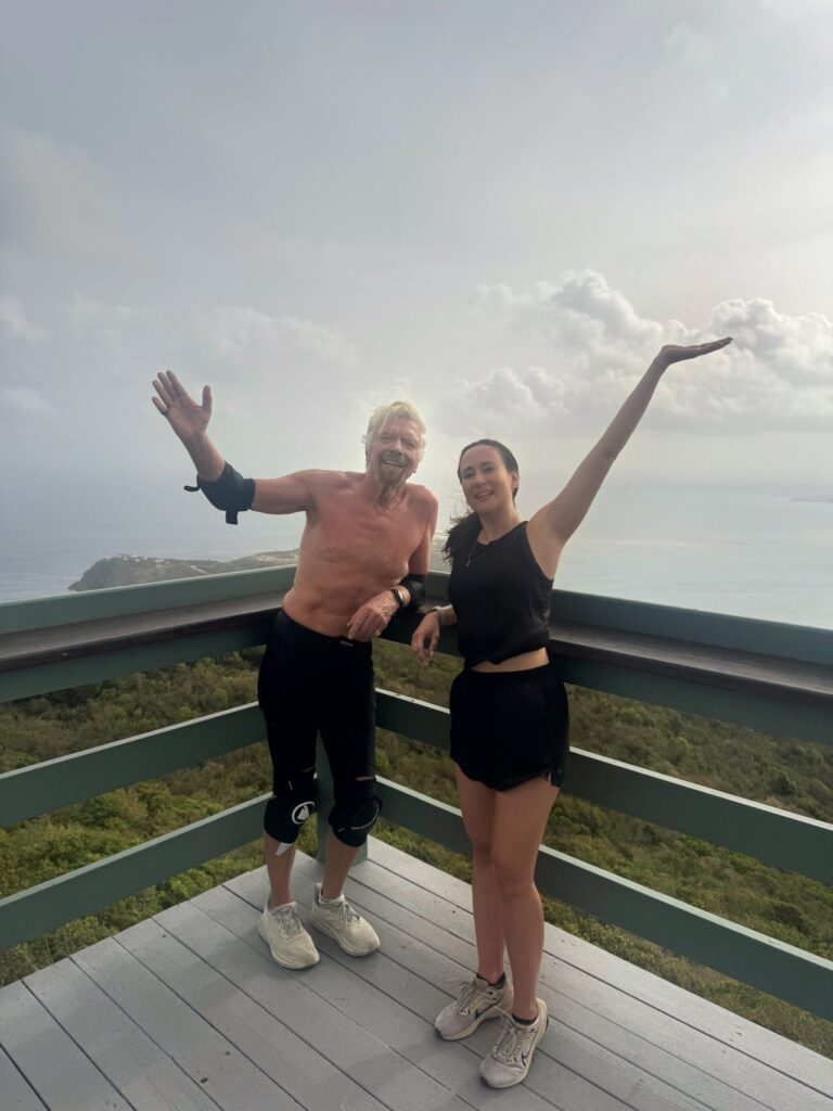 Ania Wysocka and Richard Branson posing with arms raised on an outdoor deck overlooking a tropical coastline