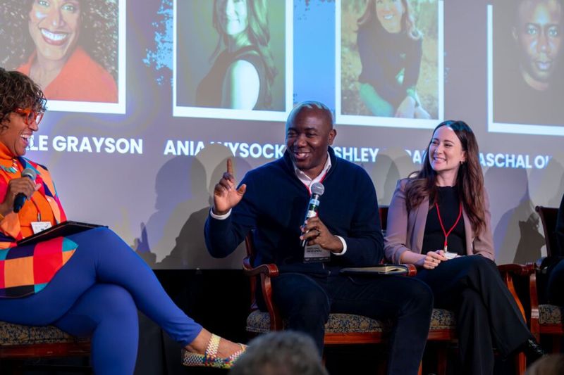 Ania Wysocka seated on a conference panel stage, listening and smiling, with a projection screen showing speaker headshots and name labels behind the panelists