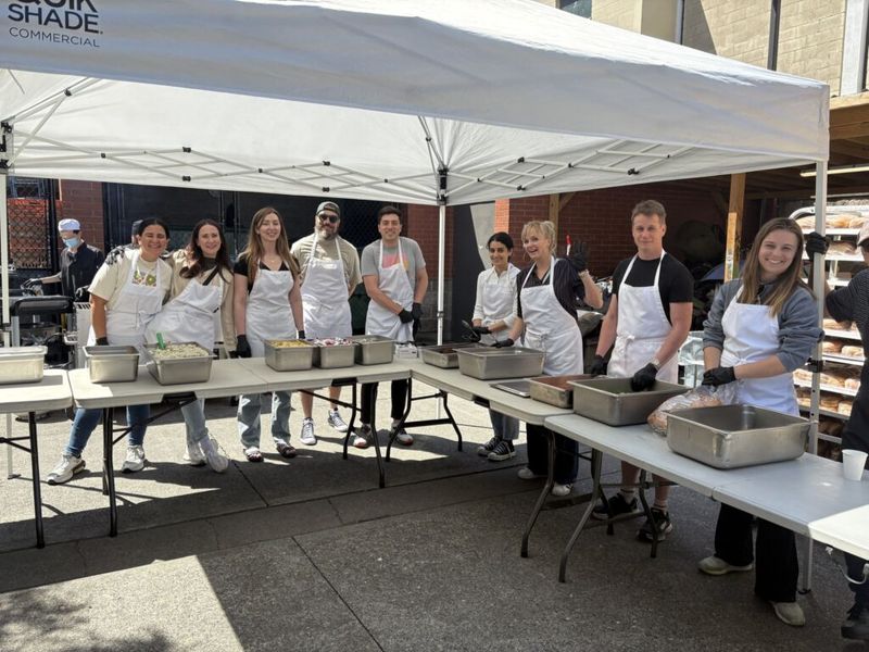 Group of volunteers in white aprons serving food at an outdoor community event under a canopy tent