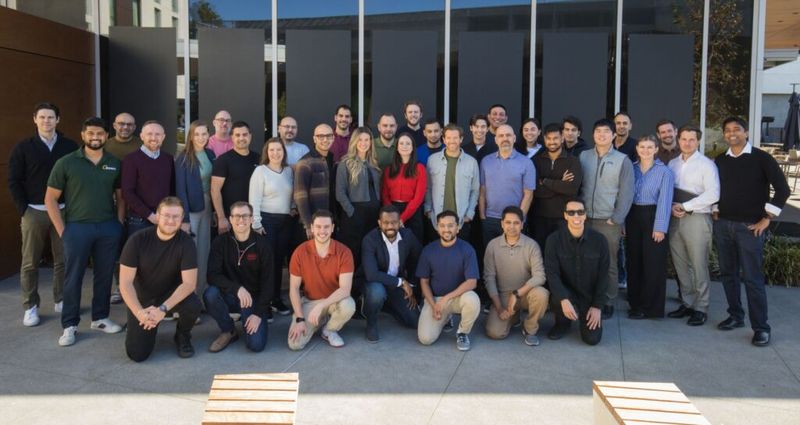 A large group of colleagues posing together outdoors in front of a modern building with dark glass panels