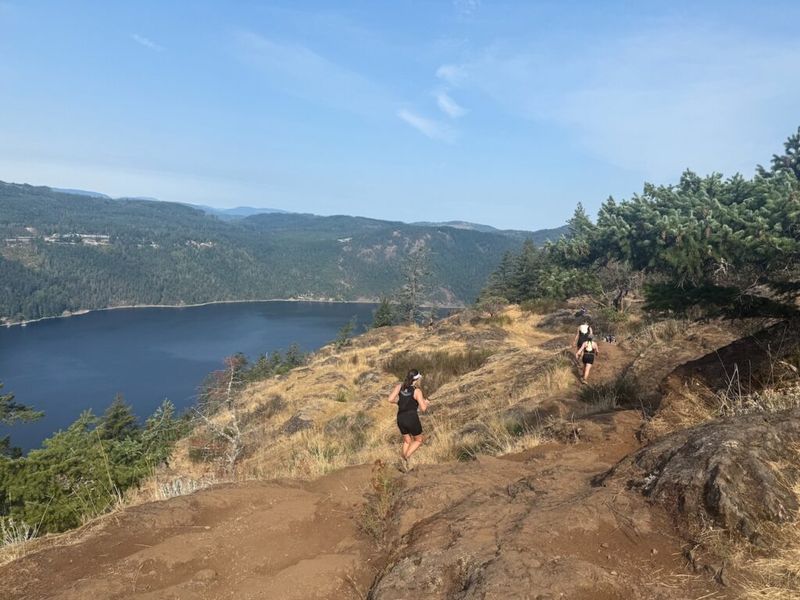 Two trail runners on a rocky ridge above a lake, surrounded by dry grass, conifers, and forested hills